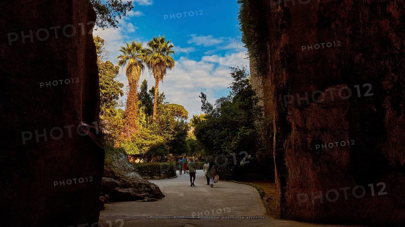 Cave Ear of Dionysus, Two people walking through a rocky passage with palm trees in the background in sunny weather, Archaeological Park Neapolis, Syracuse, Sicily, Italy