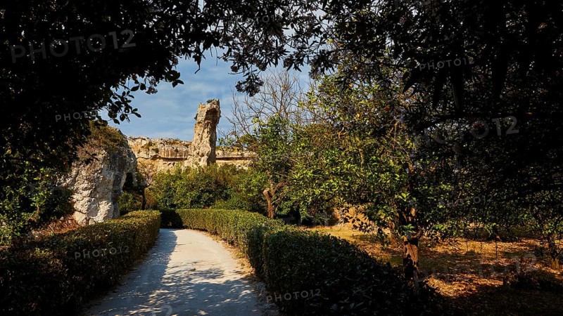 Park, A path leads through a sunlit landscape of ruins surrounded by trees and bushes, Neapolis Archaeological Park, Syracuse, Sicily, Italy