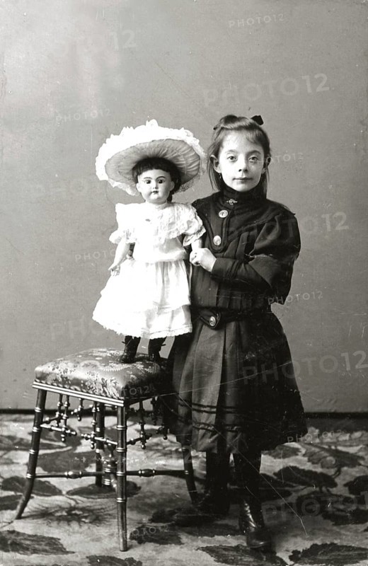 Girl doll dresses, 1930s, girl standing next to a chair carrying a doll in a dress, both in nostalgic clothes, historical photo