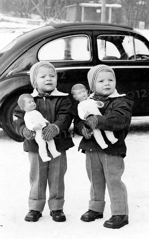 Twins, 1950s, Two children standing outside in the snow, holding dolls, car in the background, Historical photo