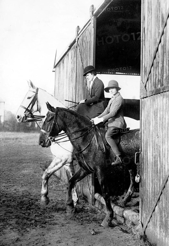 Riding horses out of a barn, 1920s, Two riders on horses coming out of a barn, wearing helmets, Historical photo