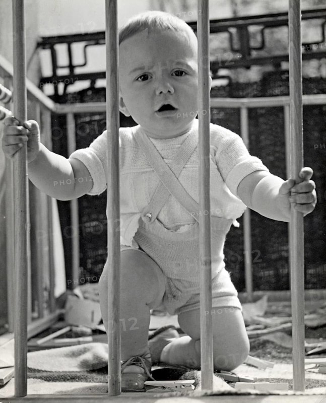 Little boy in a playpen, 1930s, Toddler kneeling in a cot and holding on to the wooden railing, historical photo