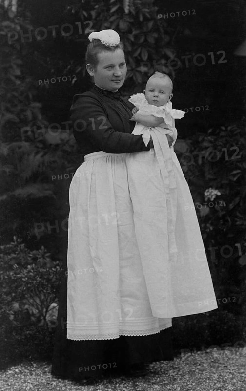 Nanny holding a baby in her arms, ca. 1910, Woman in Victorian dress holding a baby in the garden, both elegantly dressed, historical photo