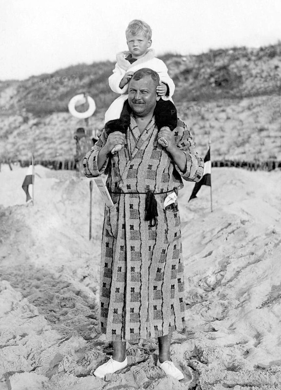 Man in bathrobe carrying child on shoulders, 1930s, A father carries his child on his shoulders on a sandy beach, Historical photo