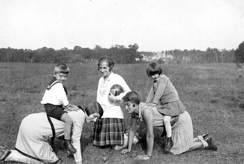 Two children riding on the backs of their mothers, 1930s, Children playing in the meadow with two adults in a cheerful atmosphere, Historical photo