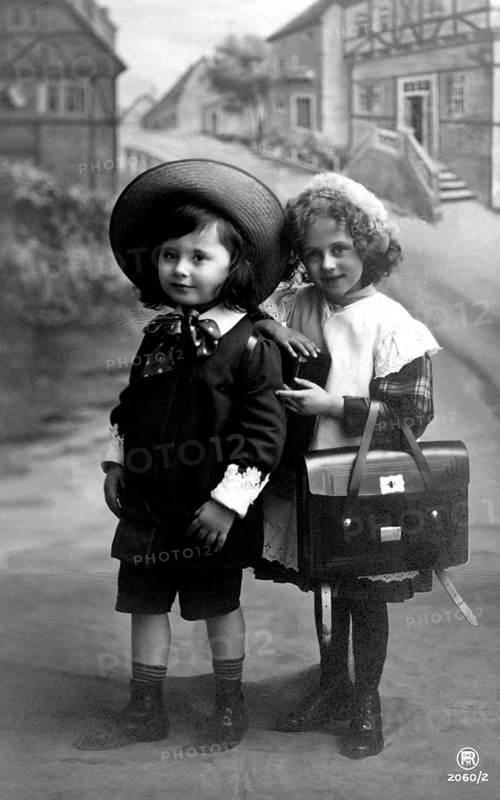 2 children in school clothes, 1910, Two children in traditional clothes with satchels, in front of a picturesque village backdrop, Historical photo