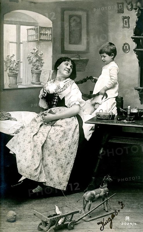 Child playing with mum's hair, 1920, A woman and a boy are inside, surrounded by toys, in a nostalgic black and white picture, Historical photo