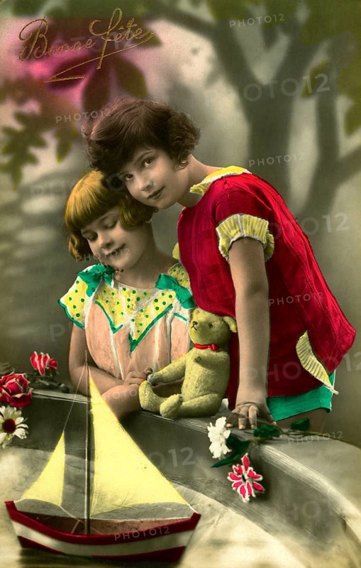 Two girls at play, 1930s, Two children in colourful clothes standing in front of a sailing boat with flowers and a stuffed bear, Historical photo