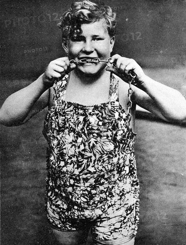 Child biting chains, 1910, A boy in a suit shows strength by pulling a chain and smiling, Historical photo