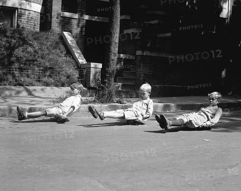 Three children sitting on skateboards, 1920s, Three boys playing on skateboards on a city street, Historical photo