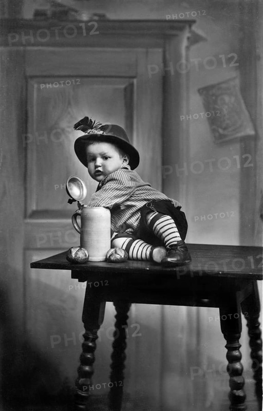 Baby traditional traditional costume beer mug, 1930s, child in traditional dress sitting on a table next to a large mug, historical photo