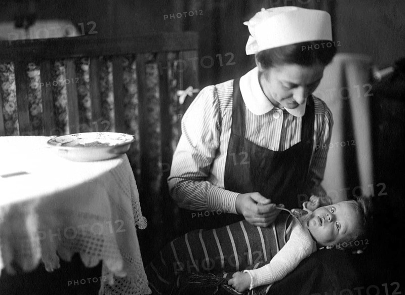 Nanny feeding child, 20s, 1920s, A woman lovingly feeds a baby in a domestic, historical setting, showing caring, Historical photo