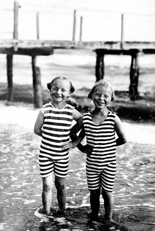 Two girls, striped swimming costume, swimwear, 1920s, Two children in striped swimming costumes on the beach, smiling in the water, wooden constructions in the background, Historical photo