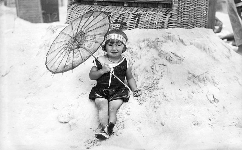 Child on the beach, 1920s, Little girl in summer clothes sitting smiling on the beach with an umbrella, Historical photo