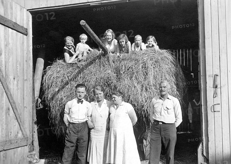 Farming family in front of the barn, ca. 1930, Family posing smiling in front of a bale of hay in an open barn, historical photo