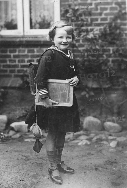 School enrolment, 1920s, A girl with a slate and school uniform stands in front of a brick wall, historical photo