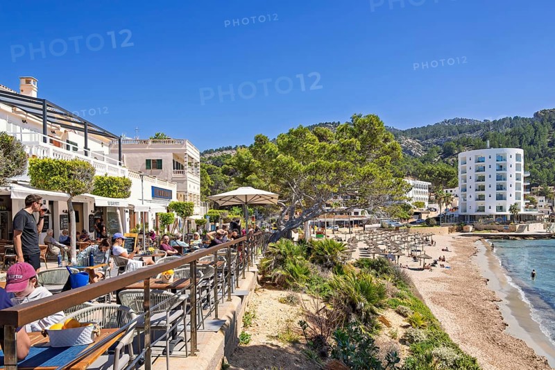Outdoors restaurant on a terrace with tourist sitting in the sunshine and looking at the seascape view in a village by the mediterranean sea, Sant Elm, Mallorca, Spain