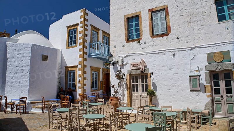 An outdoor area of a café with empty tables and chairs in front of white buildings on a sunny day, Platia Lesvias, Chora, UNESCO World Heritage Site 1999, Patmos, Dodecanese, Greek Islands, Greece