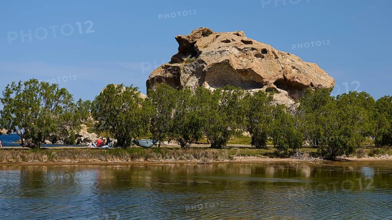 Rock rises behind a row of trees on the lakeshore under a clear sky, Rock of Kalikatsou, Seasonal Salt Lake, Patmos, Dodecanese, Greek Islands, Greece