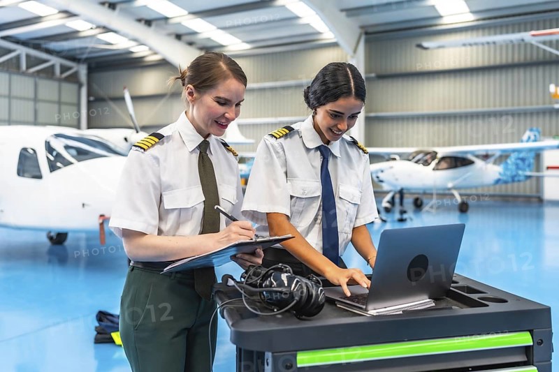 Two female pilots in uniform working on a laptop and clipboard in a spacious aircraft hangar, surrounded by small planes