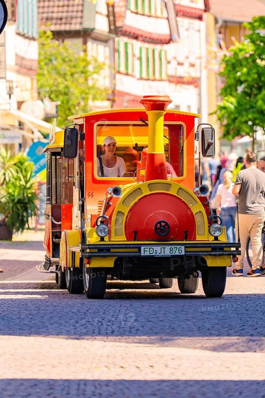 Small colourful train runs through a lively old town with cheerful people, Calw station festival, Black Forest, Germany