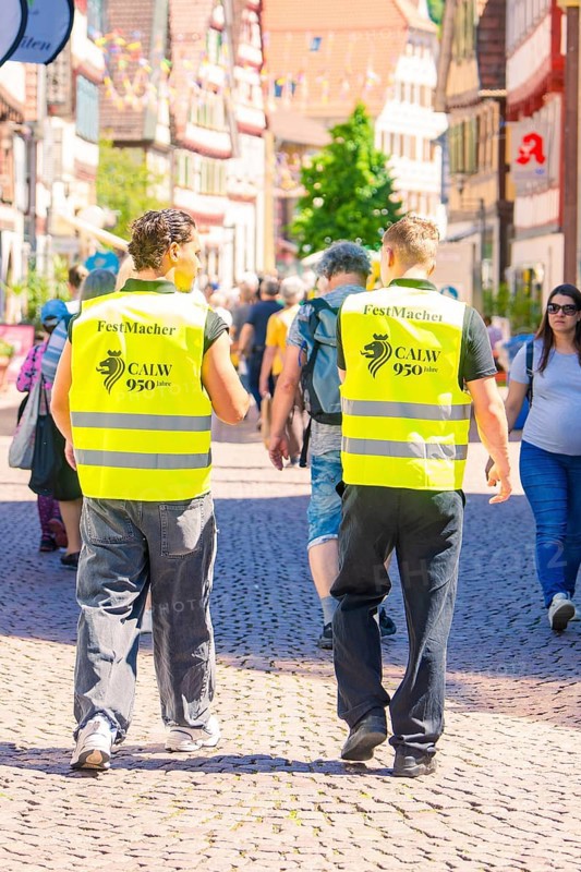 Two men in yellow waistcoats walk along a busy, sunny street, Calw railway station festival, Black Forest, Germany