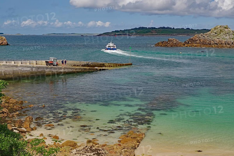 A boat approaches the harbour on clear blue water surrounded by rocks and islands, Ferry, St Agnes, Isles of Scilly, Cornwall, England, United Kingdom
