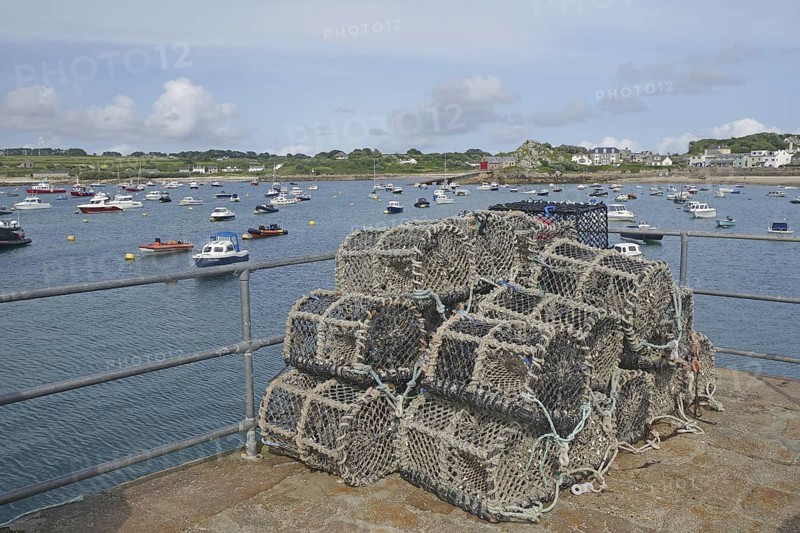 A harbour with stacked lobster pots, many boats on the water and a coastline in the background, Isles of Scilly, Cornwall, England, Great Britain