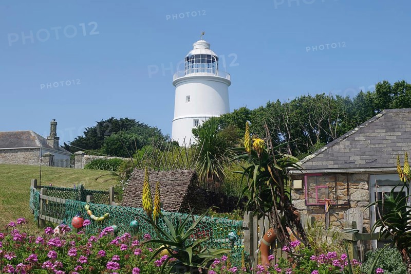 A lighthouse next to a sunny garden with blooming flowers and stone houses under a blue sky, St Agnes, Isles of Scilly, Cornwall, England, United Kingdom