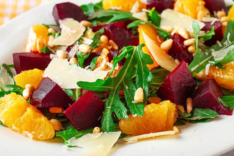 A salad made with chunks of beet, orange slices, and arugula. It is topped with cheese shavings and pine nuts, placed on a white plate with a striped napkin beside it