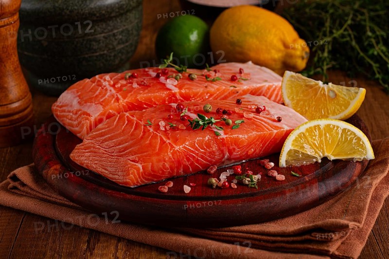 Two salmon fillets rest on a wooden platter. The fillets are garnished with herbs and spices. Lemon and lime slices are nearby. This setup is ready for preparation in a kitchen