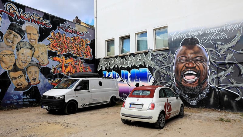 Murals of the local graffiti scene. The portrait of Keno Veith is particularly distinctive, with the saying Wenn Löppt dann Löpps, Munter. Another mural shows Dieter Bohlen together with faces that allude to the Bohlen's Right Sons theme, preceded by a Fiat 500 convertible and a VW Transport, Oldenburg, Lower Saxony, Germany
