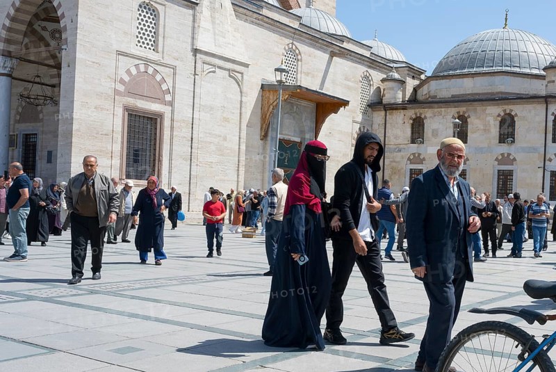 People wearing traditional clothes walking on a sunny square in front of historic buildings with domes, woman wearing burqa, Konya Sultan Selim Camii, Konya, Central Anatolia, Turkey