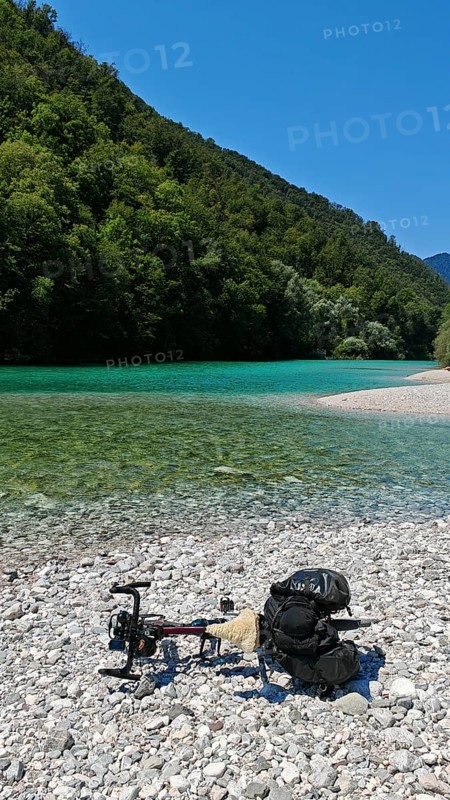 A bicycle is lying on a pebble beach by a river with clear water and wooded banks, Soca Valley, Slovenia