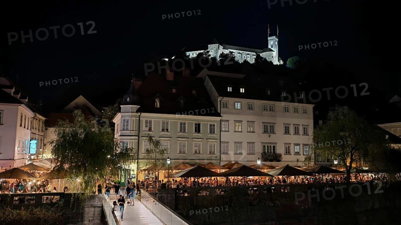 Castle illuminated at night surrounded by illuminated restaurants and visitors in an urban setting, Ljubljana, Slovenia