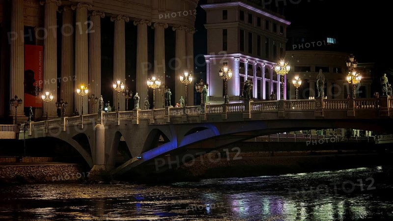 Illuminated bridge over a river at night with urban architecture in the background, Skopje, North Macedonia