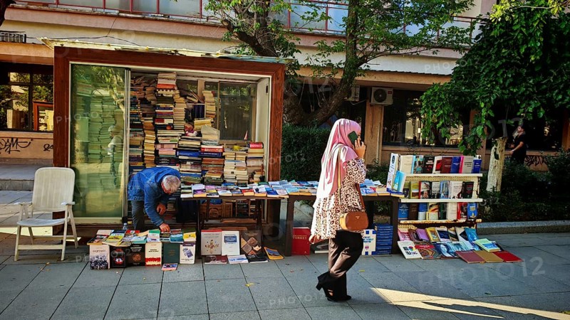 Book stand on a street surrounded by people and green trees, idyllic street scene with woman talking on the phone, Pristina, Kosovo
