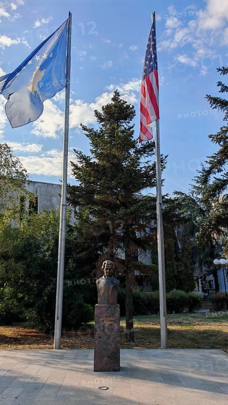 Bust statue of Madeleine Albright outdoors with two USA and Kosovo flags surrounded by tall trees, blue sky in the background, Pristina, Kosovo