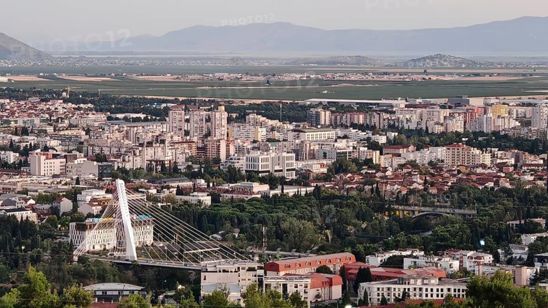 A city surrounded by nature with trees in the foreground and mountains in the background, illuminated by a warm sunset, looking out from a hill on the outskirts of Podgorica, Montenegro