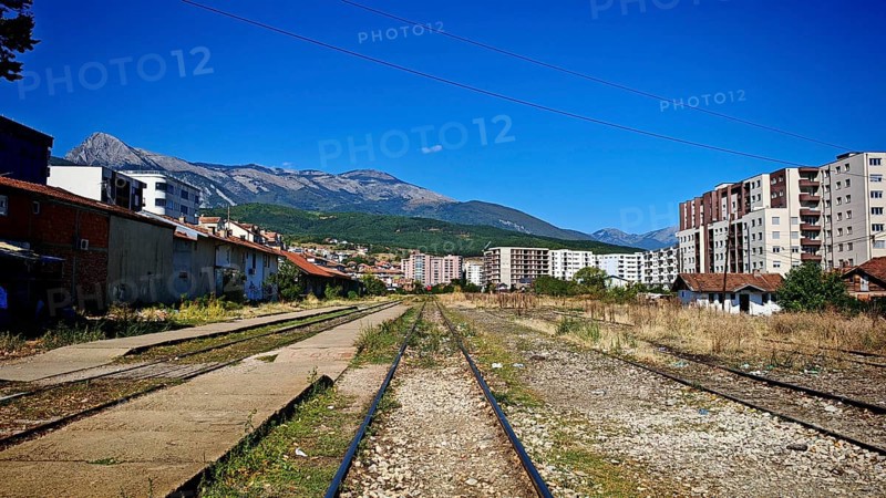 Overgrown train tracks run through a town at the foot of mountains on a sunny day, Kosovo