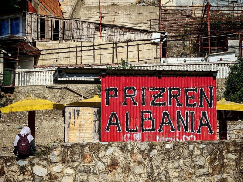 Red sign with the inscription Prizren Albania in front of building and road, Prizren, Kosovo