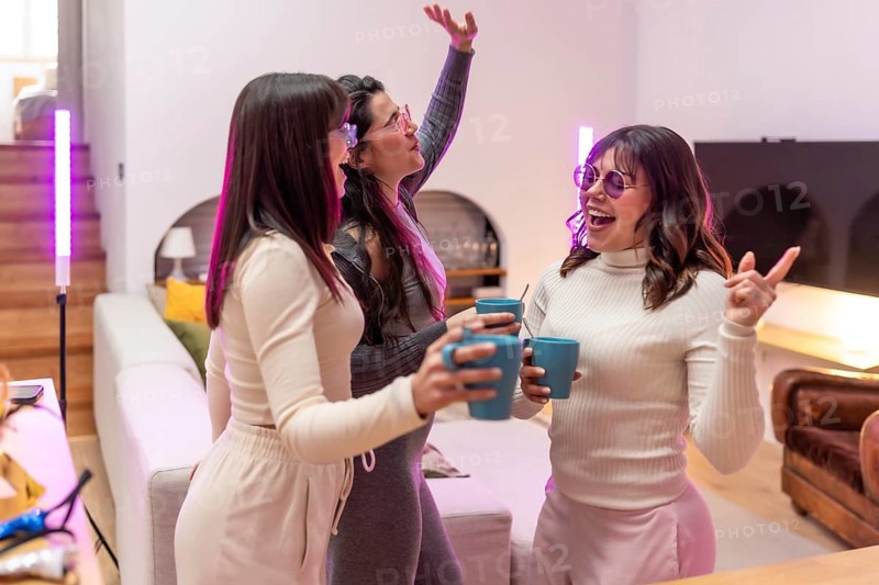 Three young women laughing and celebrating with drinks in a cozy apartment living room, purple pink party lighting creating a vibrant, candid moment of friendship and joy