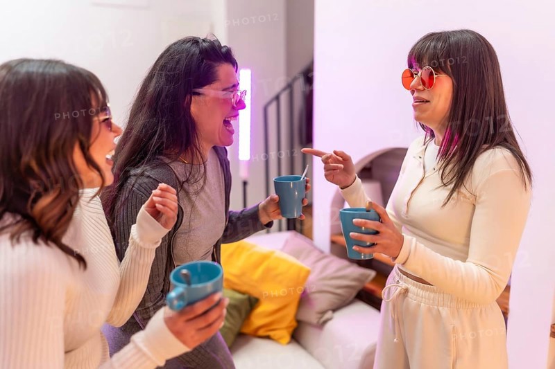 Three young women laughing and chatting over hot drinks in a cozy apartment, enjoying a relaxed evening of friendship, warmth, and casual conversation