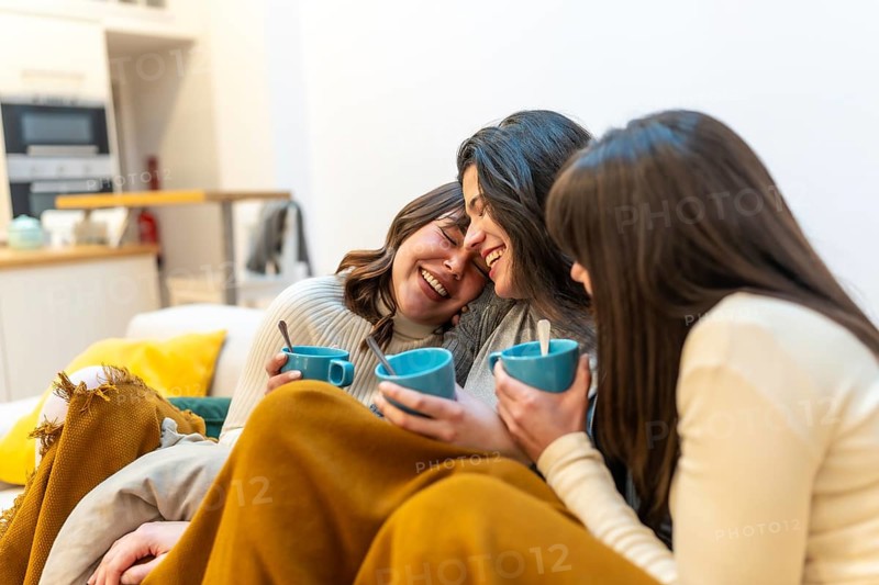 Three young women relaxing on a sofa at home, wrapped in a blanket, laughing and sharing warm drinks in a cozy, casual living room moment of friendship and comfort