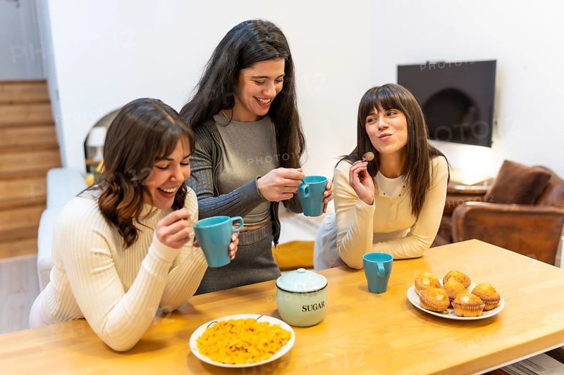 Three young women smiling and laughing, holding cups and spoons, gathering around a wooden kitchen counter with muffins and cereal for a friendly morning social event