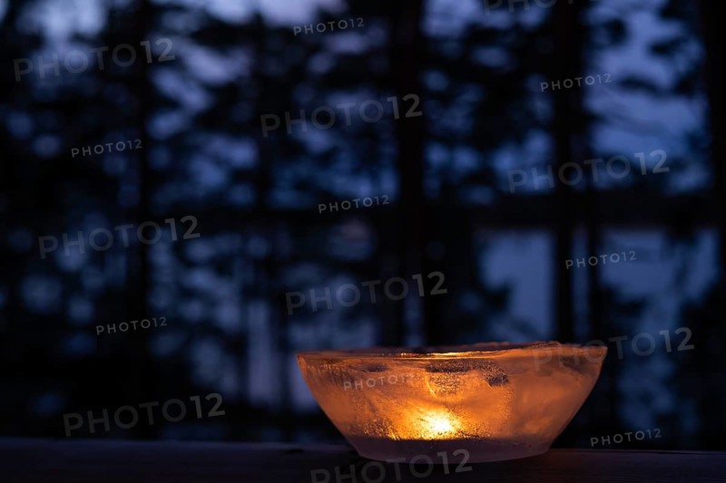 Ice bowl illuminated by burning tea light stands on balcony balustrade, forest at the onset of twilight, winter, Finland