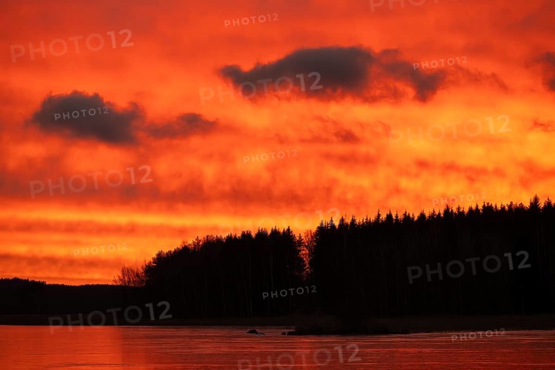 A dramatic sunset over a still, frozen lake and forest with orange-red hues in a cloudy sky, Finland