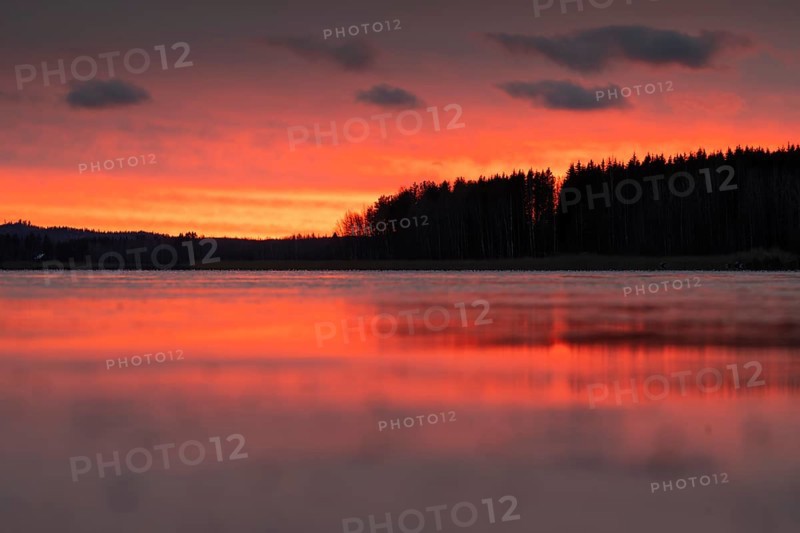 A calm sunset sky with orange-red-yellow hues over a quiet lake and dark forest, Finland