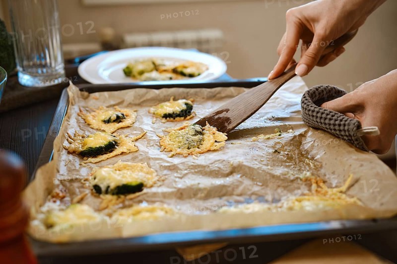 Unrecognizable woman transfers roasted smashed broccoli with cheese from baking sheet to plate using wooden spatula, close-up view