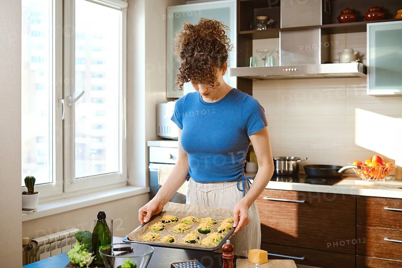 A person is in a kitchen holding a tray of smashed broccoli with cheese. Green broccoli is on top of a mix of cheese. Woman is going to put baking sheet in the oven